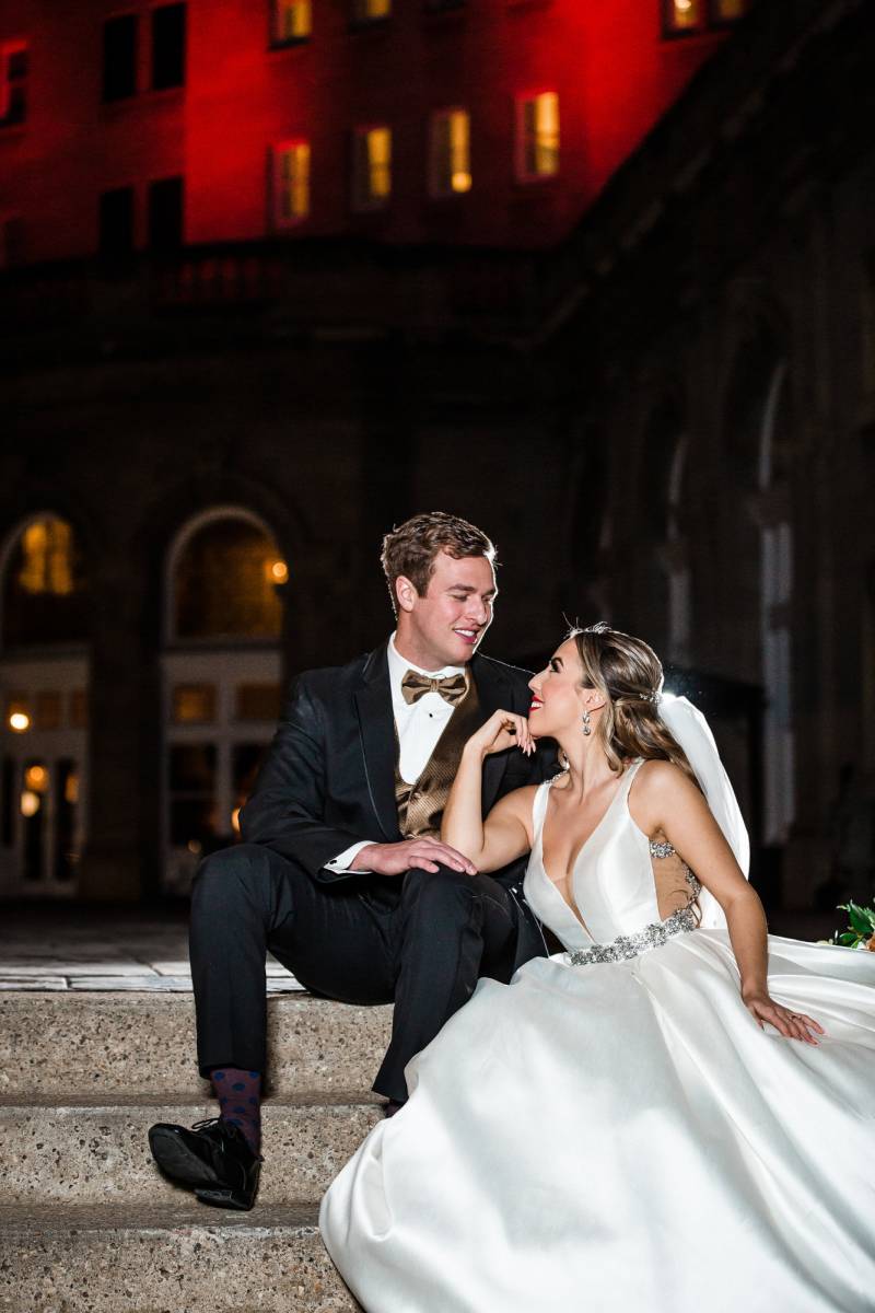 Bride wearing white dress and groom sitting on concrete stairs outside 