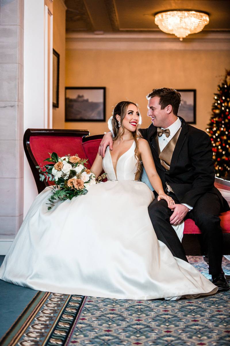 Groom and Bride in white bodice dress holding peach and white bouquet sitting on red couch 