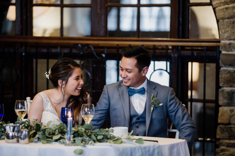 Bride and groom sit laughing sitting at white table with greenery 