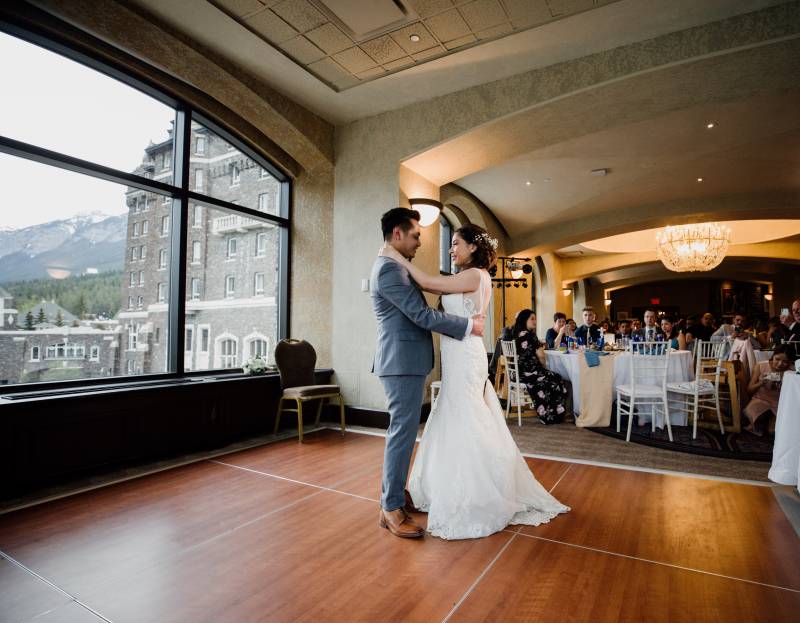 Bride and groom dance on wooden dancefloor while guests sit in the background 