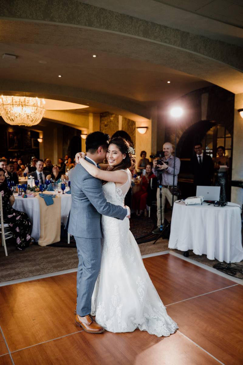 Bride and groom embrace smiling on dance floor while guests sit in the background 