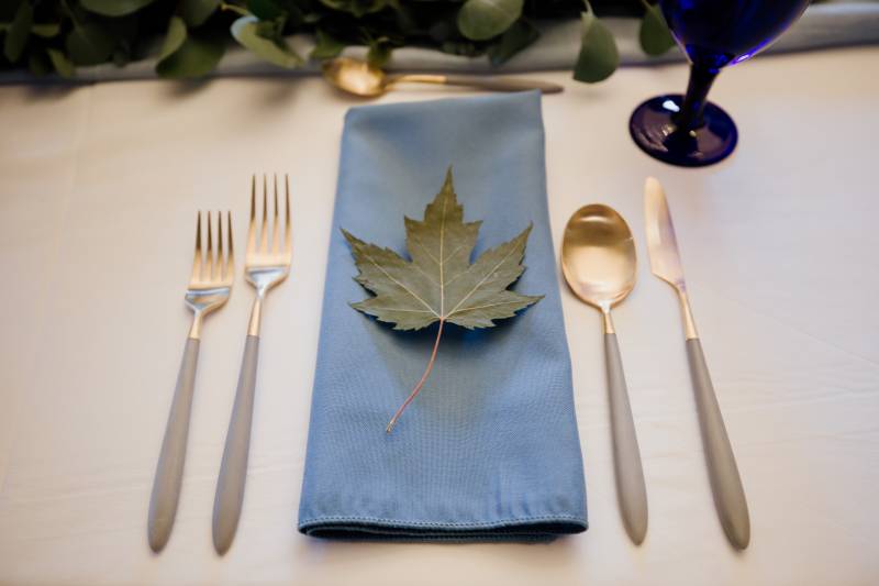 Blue table cloth with green leaf on top and gold cutlery around 