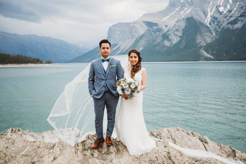Bride and groom stand on rocky slope in front of lake and mountains veil blowing behind  