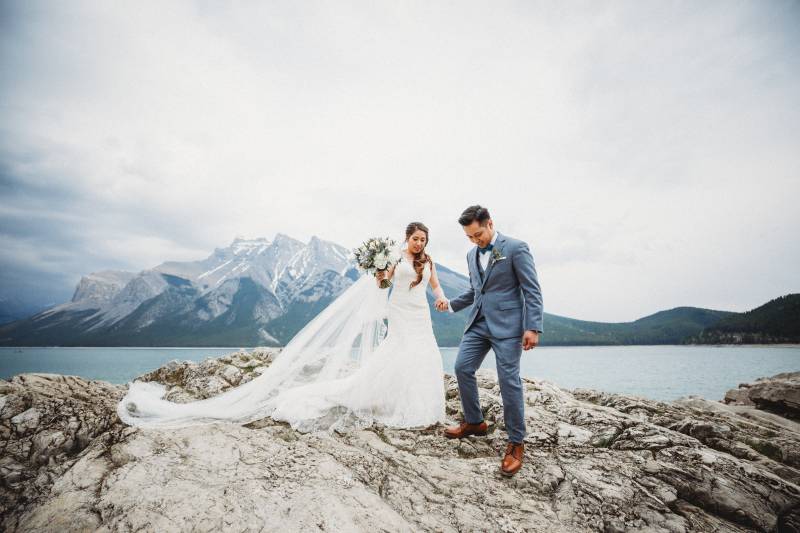 Bride and groom step down from rocky slope holding hands with veil flowing be hid 