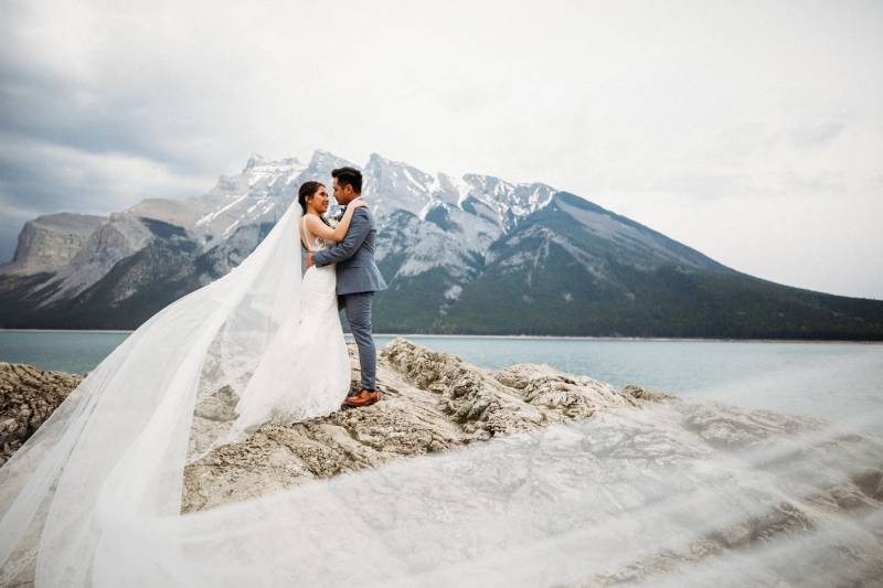 Bride and groom stand embraced on rocky ledge with long white veil flowing in wind 