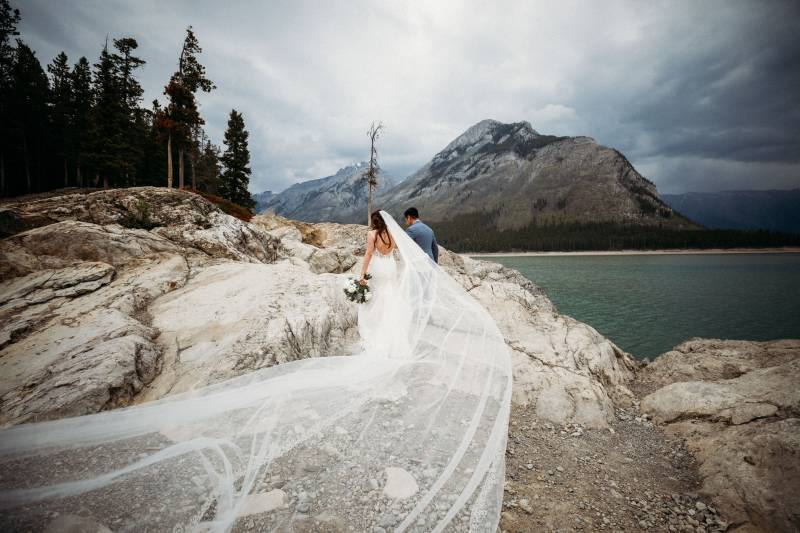 Bride and groom walk up rocky slope with long white veil flowing in wind behind 