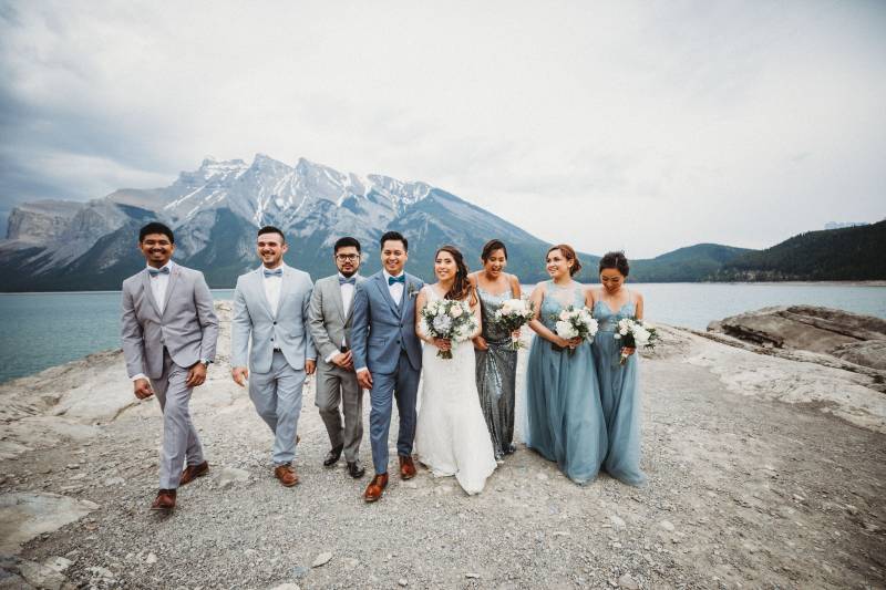 Bride and groom walk between bridesmaids and groomsmen on rocky ledge overlooking mountains and lake 