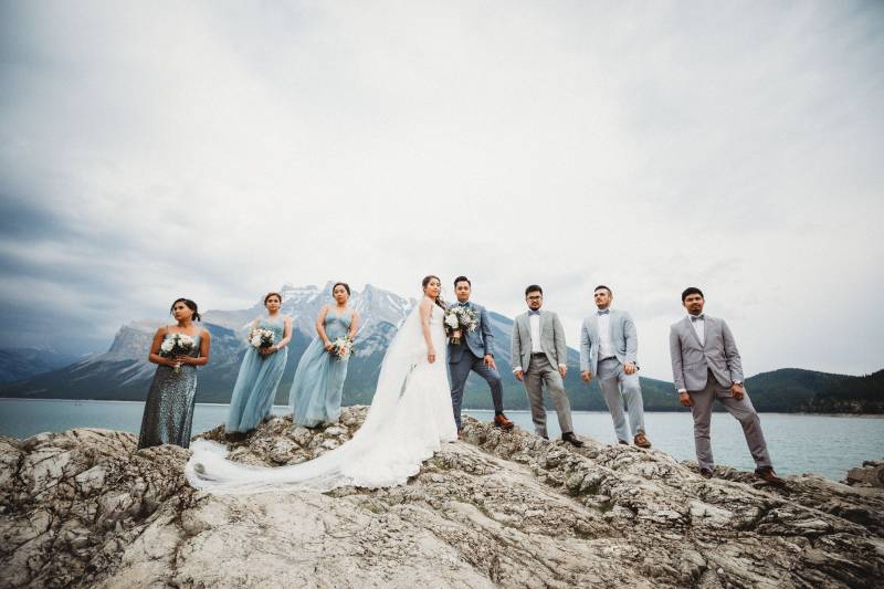 Bride and groom stand between bridesmaids and groomsmen on rocky ledge overlooking lake and mountains 