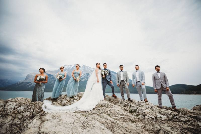 Bride and groom stand between bridesmaids and groomsmen on rocky ledge overlooking lake and mountains 
