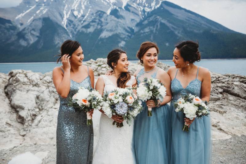 bride stands between bridesmaids smiling holding white bouquets backing mountains and lake 
