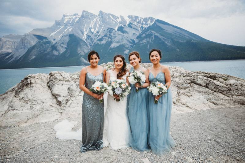 bride stands between bridesmaids holding white bouquets backing mountains and lake 