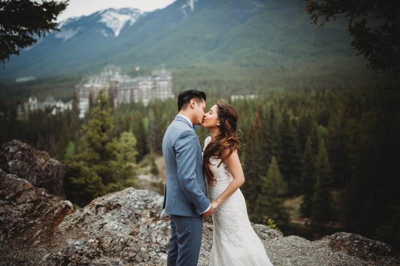 Bride and groom kiss holding hands overlooking forest and mountains 