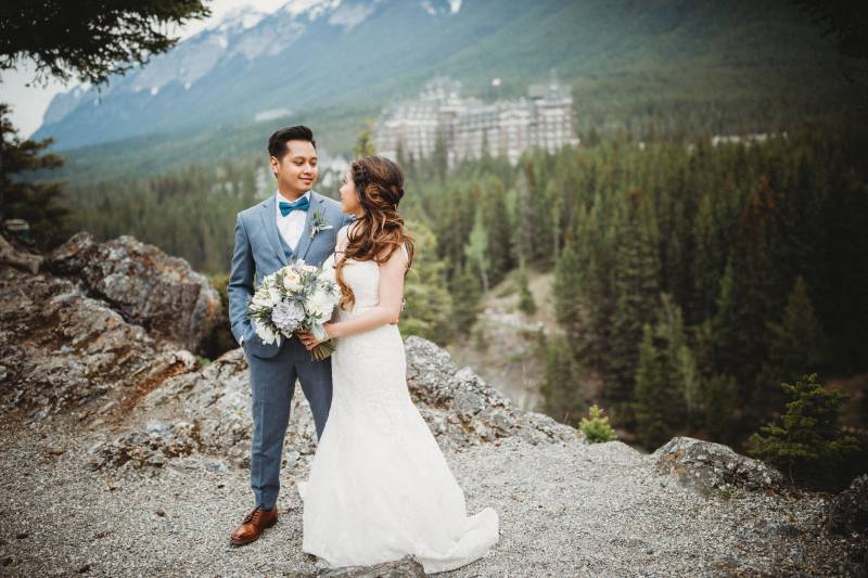 Bride and groom stand looking at each other on rocky ledge in front of forest and mountains 