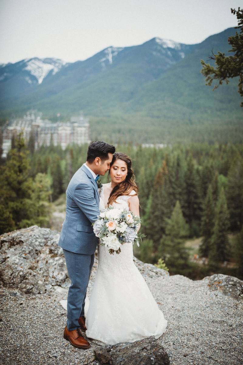 Bride stands holding white and blush bouquet while groom embraces from side on rocky ledge backing mountains and forest 