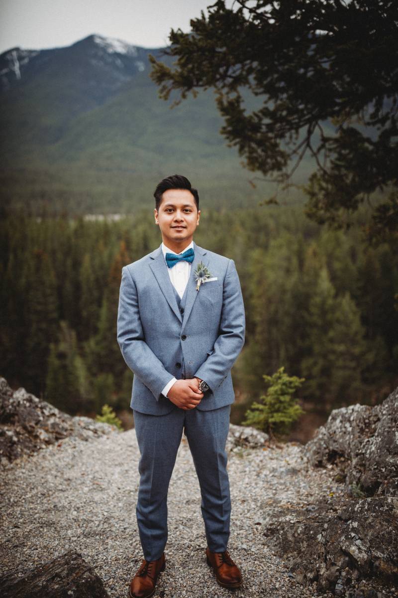 Groom stands in light blue suit hands crossed on rocky ledge backing forest and mountains 