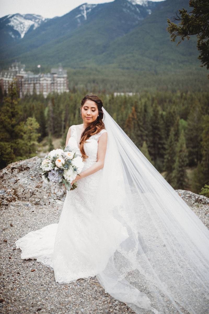 Bride sits in large white dress and veil flowing in wind holding blush and white bouquet overlooking forest and mountains 