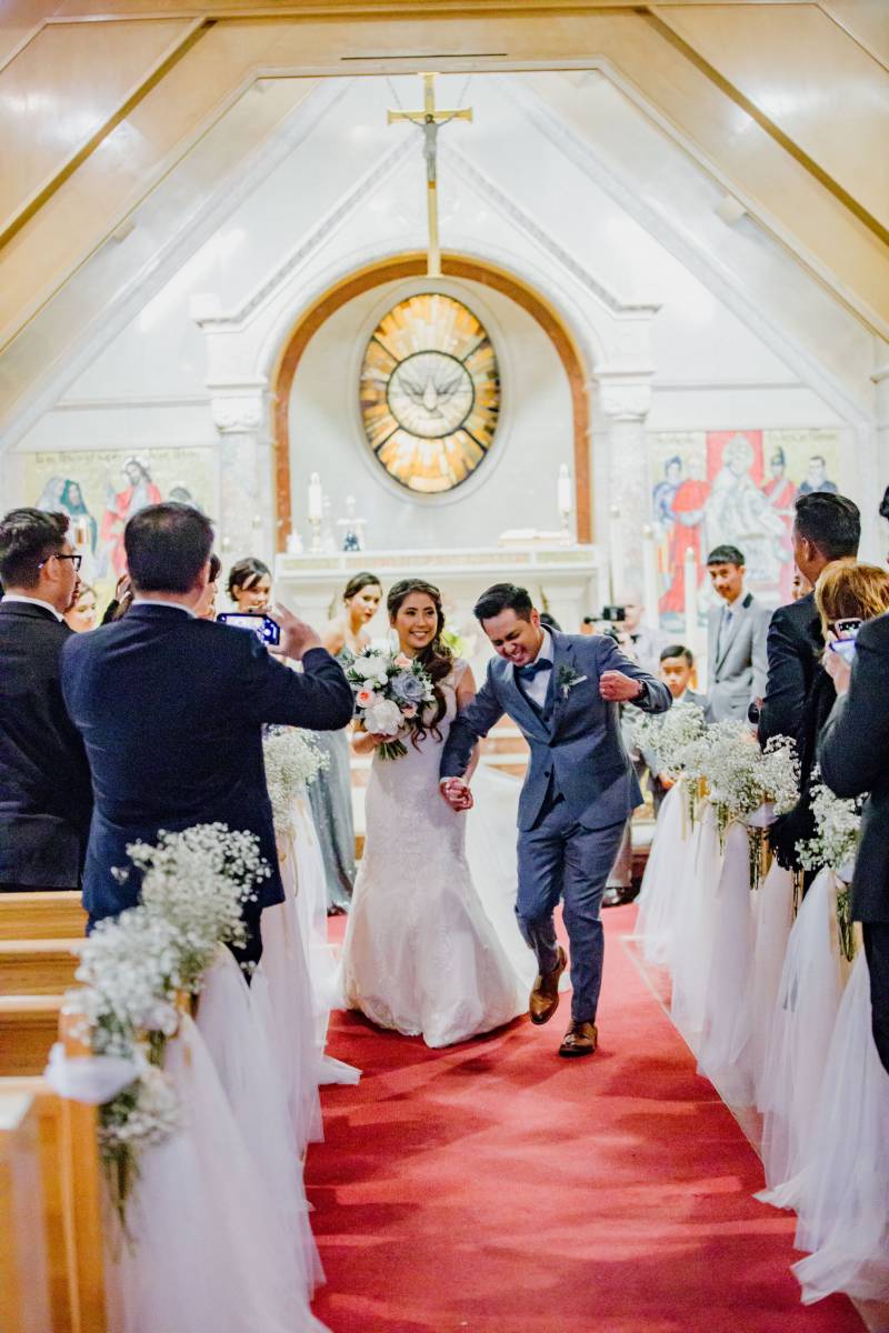 Groom walks down aisle in hands with bride smiling shaking fist excitedly while guests watch and film