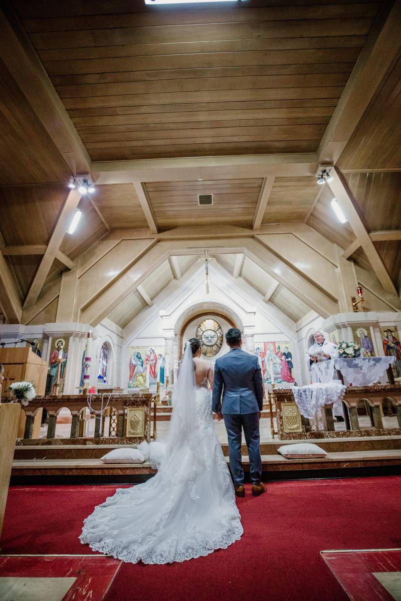 Bride in large white lace dress stands beside groom in blue suit facing front of church 