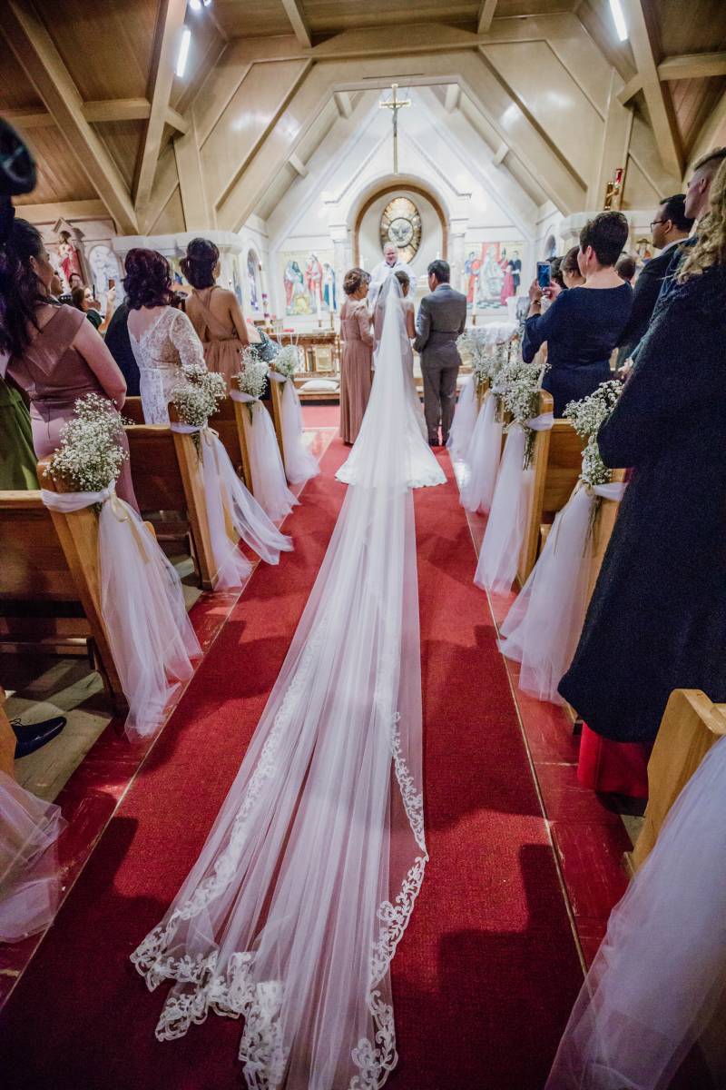 Bride walks down aisle with long white lace veil dragging down length of aisle 