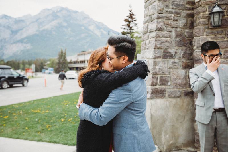 Man in blue suit hugs woman inn black coat beside stone building 