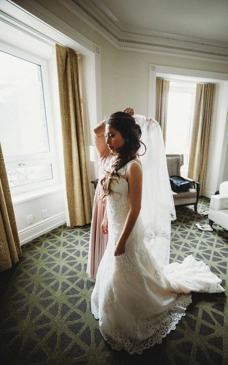 Bride stands in white lace dress while woman applies white veil to hair 