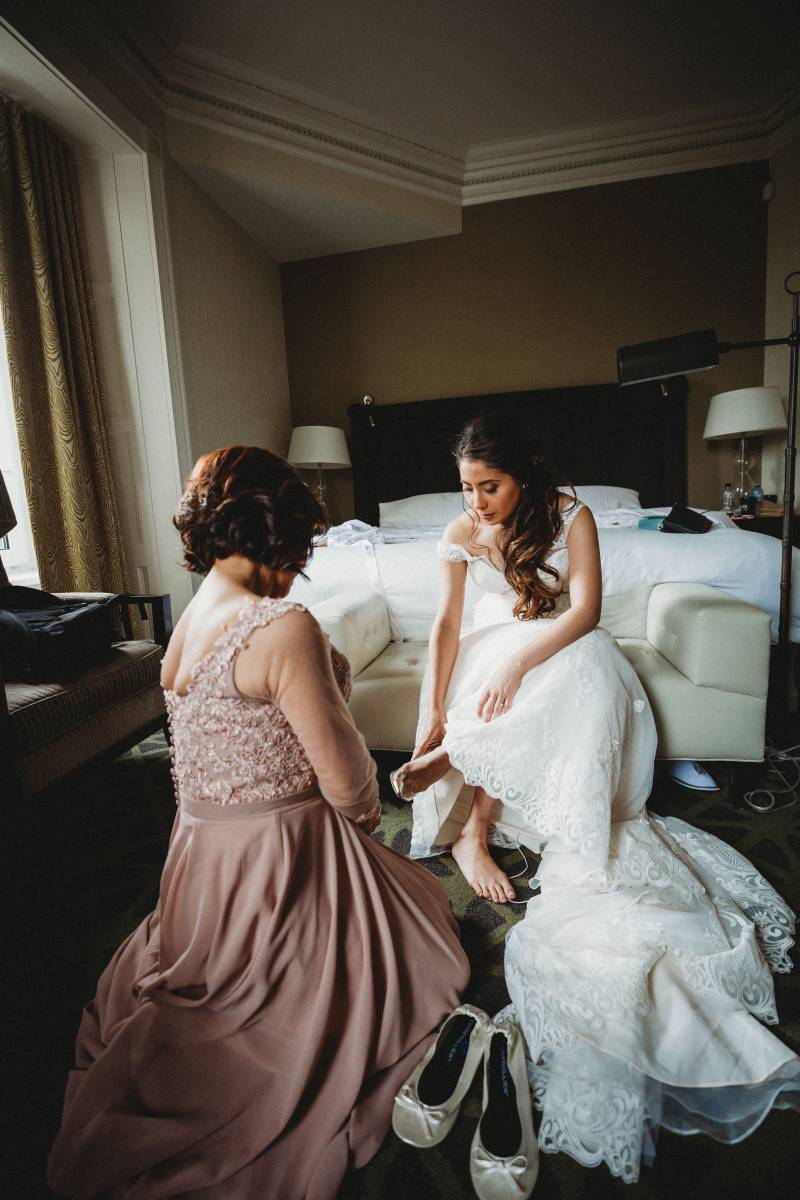 Bride sits in white lace dress at the foot of bed putting on shoe while woman sits in front 