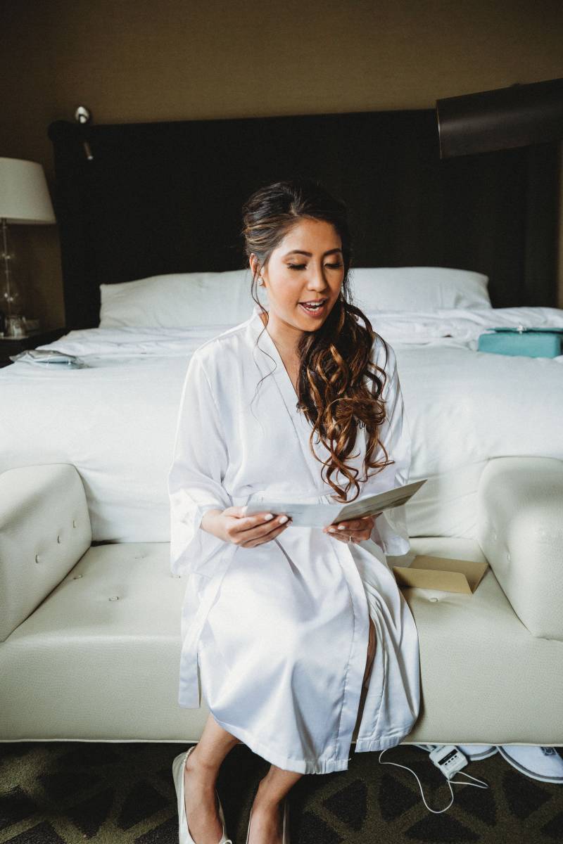 Bride sits in white robe at foot of bed reading card 