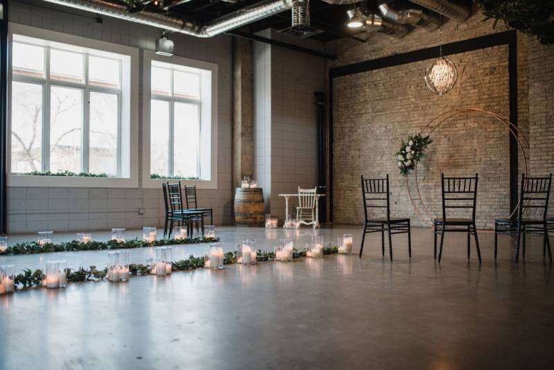 Small industrial brick and concrete room with pathway of candles and green with copper wedding arch