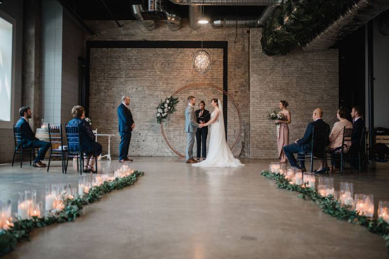 Man and woman hold hands in front of officiant and copper wedding arch and chandelier with guests in front