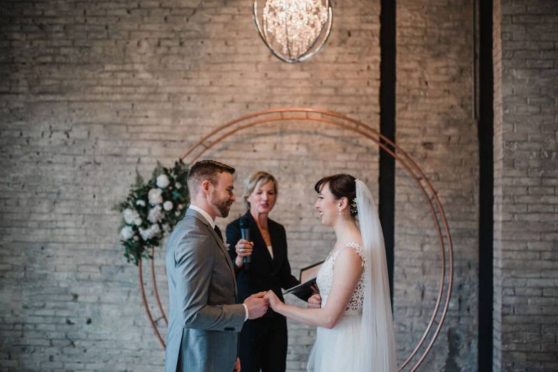 Man and woman hold hands in front of officiant and copper wedding arch and chandelier 