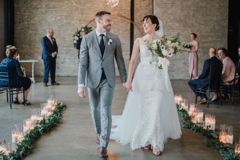 Man and woman smile together while walking down aisle holding hands between candle and vine path
