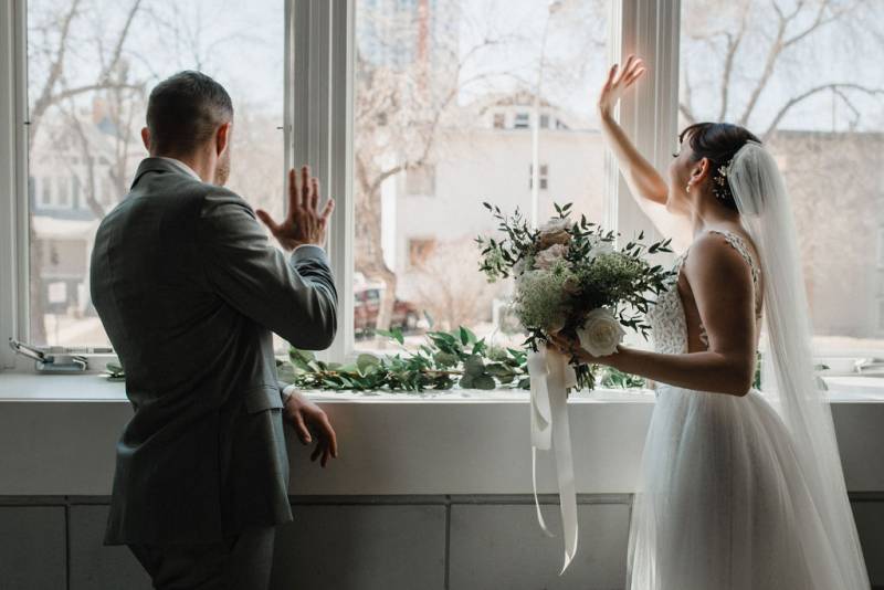 Bride and groom wave through a window decorated with green foliage 