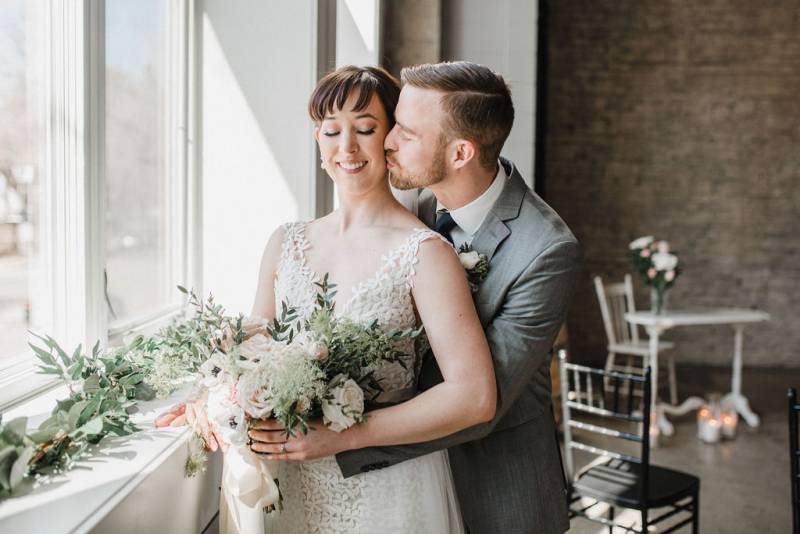 Man embraces woman wearing white lace dress from behind while holding a white bouquet 