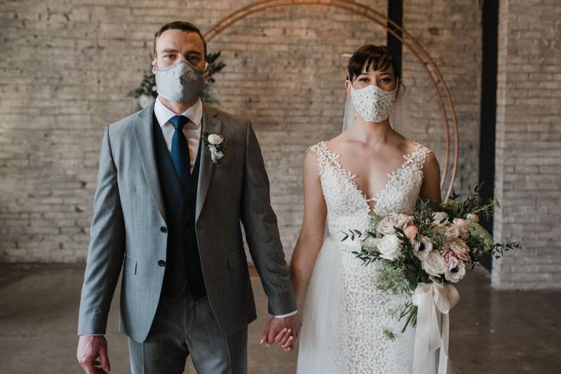 Man and woman hold hands while wearing face masks standing in front of copper wedding arch 
