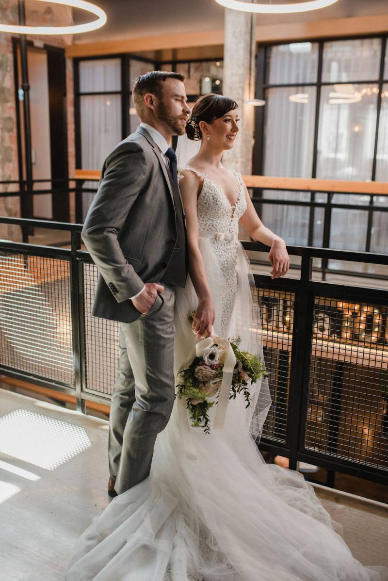 Bride and groom lean against black railing under glowing ring chandeliers 