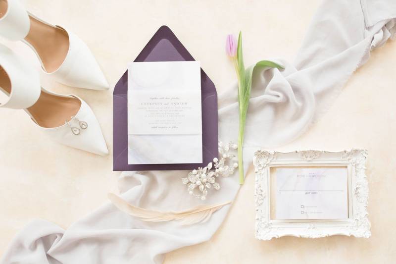 Wedding invitation flat lay on purple envelope on white silk fabric beside white heels