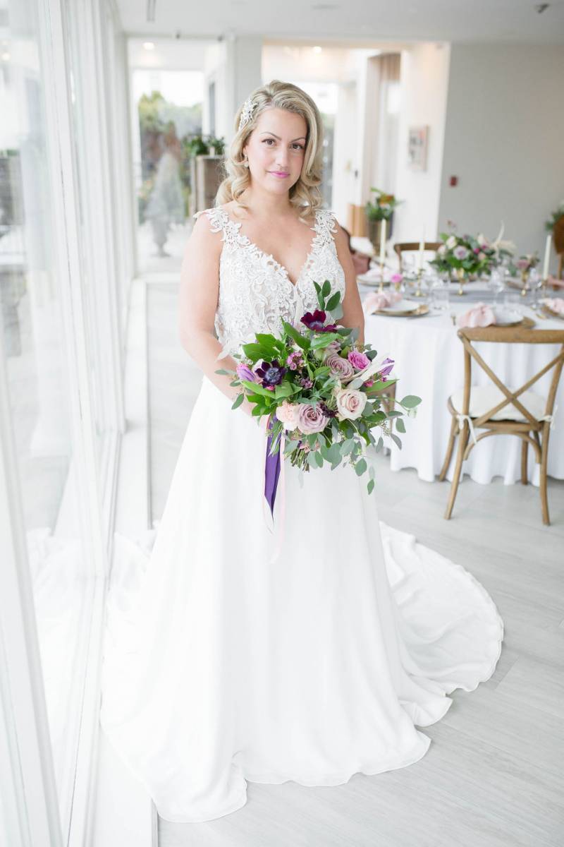 Bride in white lace dress holding bouquet with purple ribbon beside window
