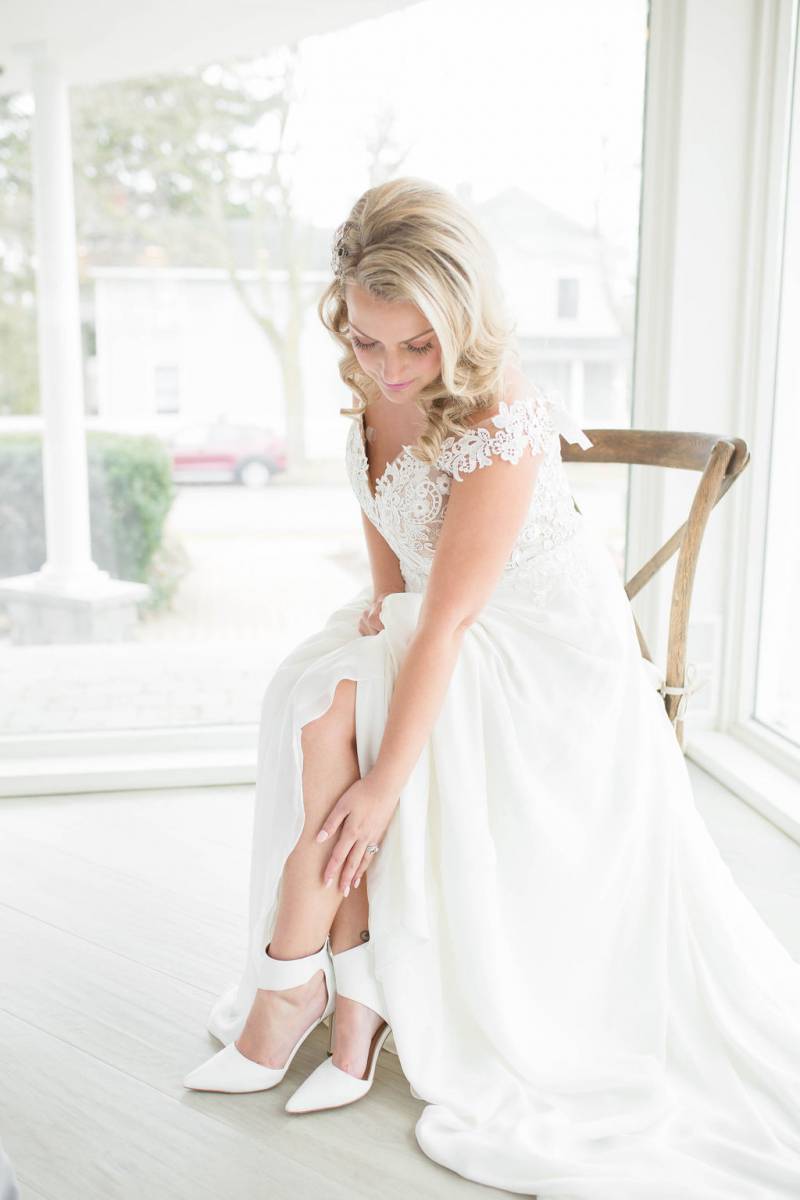 Bride sitting in white lace dress reaches down to ankles showing white heels