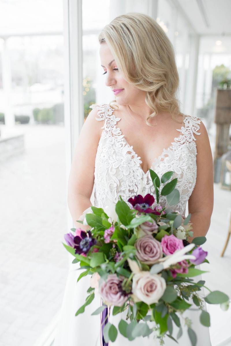 Bride in white lace dress holding bouquet with purple ribbon beside window