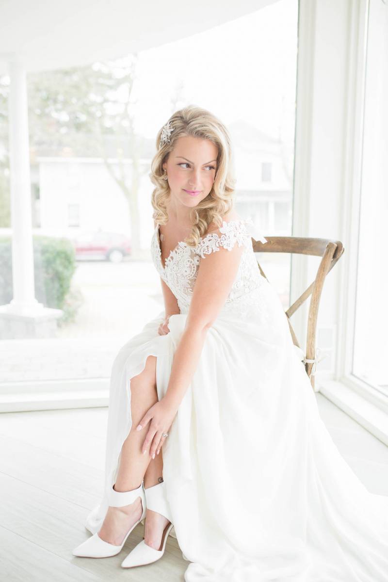 Bride sitting in white lace dress reaches down to ankles showing white heels