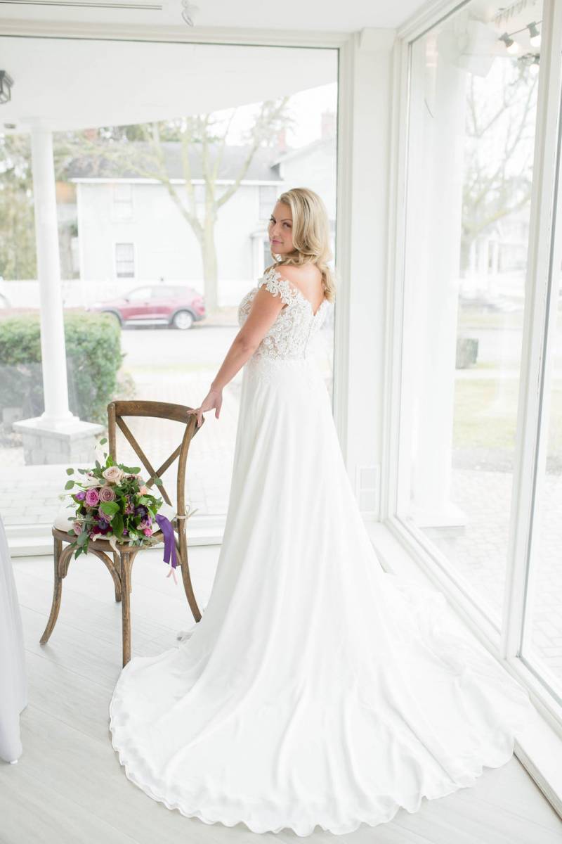 Bride standing in white lace dress leaning on wooden chair looking backwards in front of window