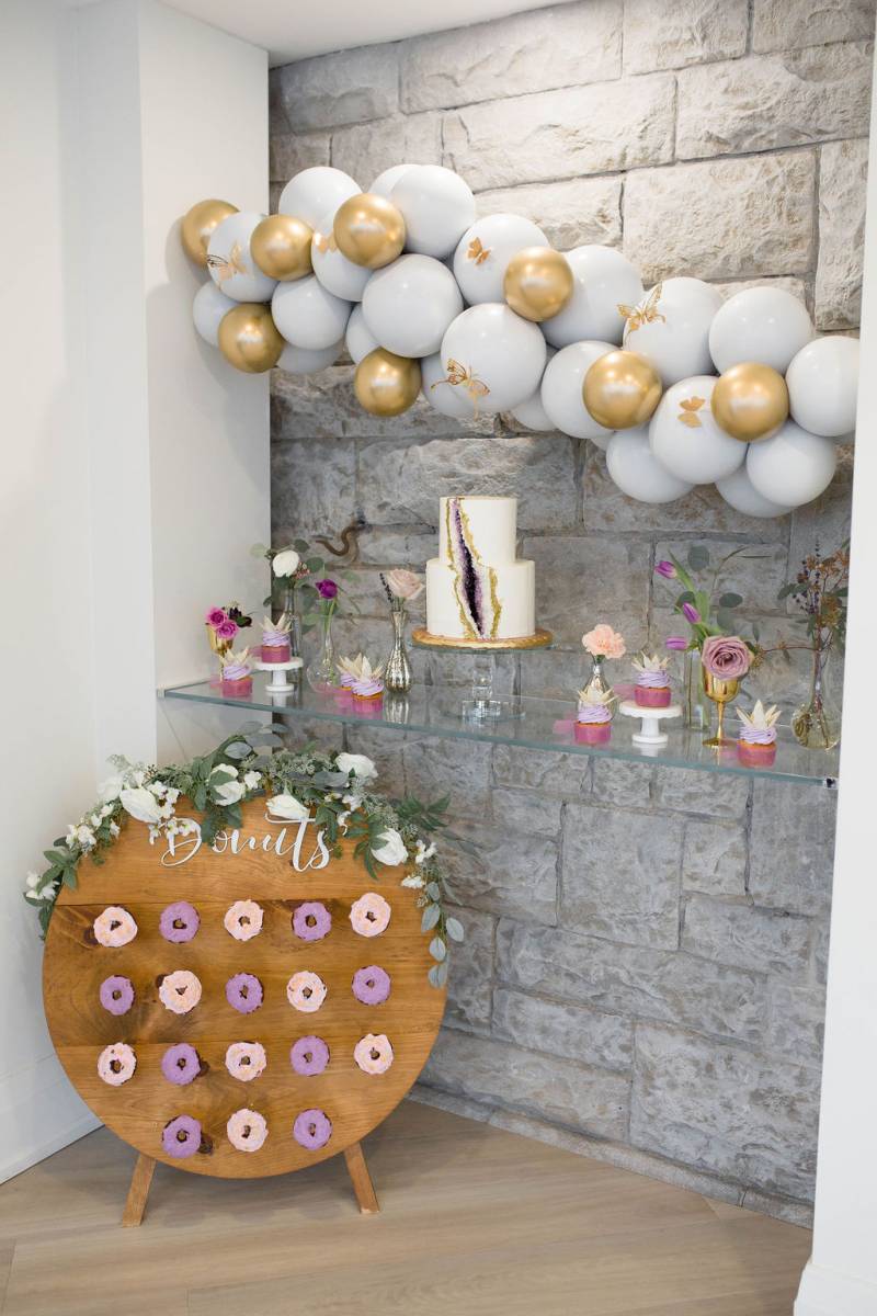 Wedding cake with purple rock slit under baby blue and gold accented balloon arch over wooden doughnut wall