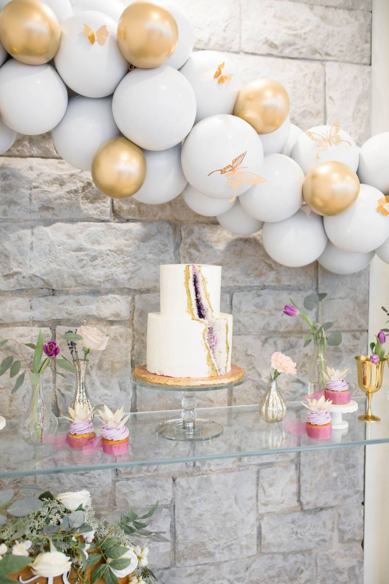 White wedding cake with purple and gold slice underneath baby blue and gold balloon arch  