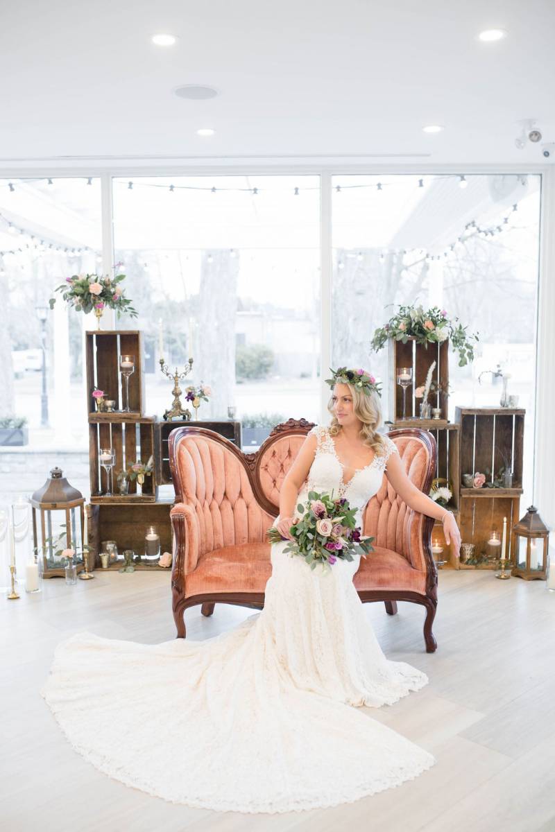 Bride leaning on salmon antique chair in large white dress and veil with flower crown holding bouquet 