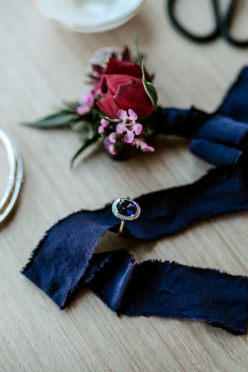 Sapphire ring on blue ribbon beside red boutonniere  