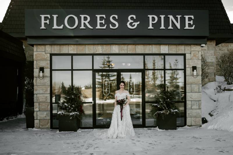 Woman in white dress holding bouquet outside snowy stone building with black roof