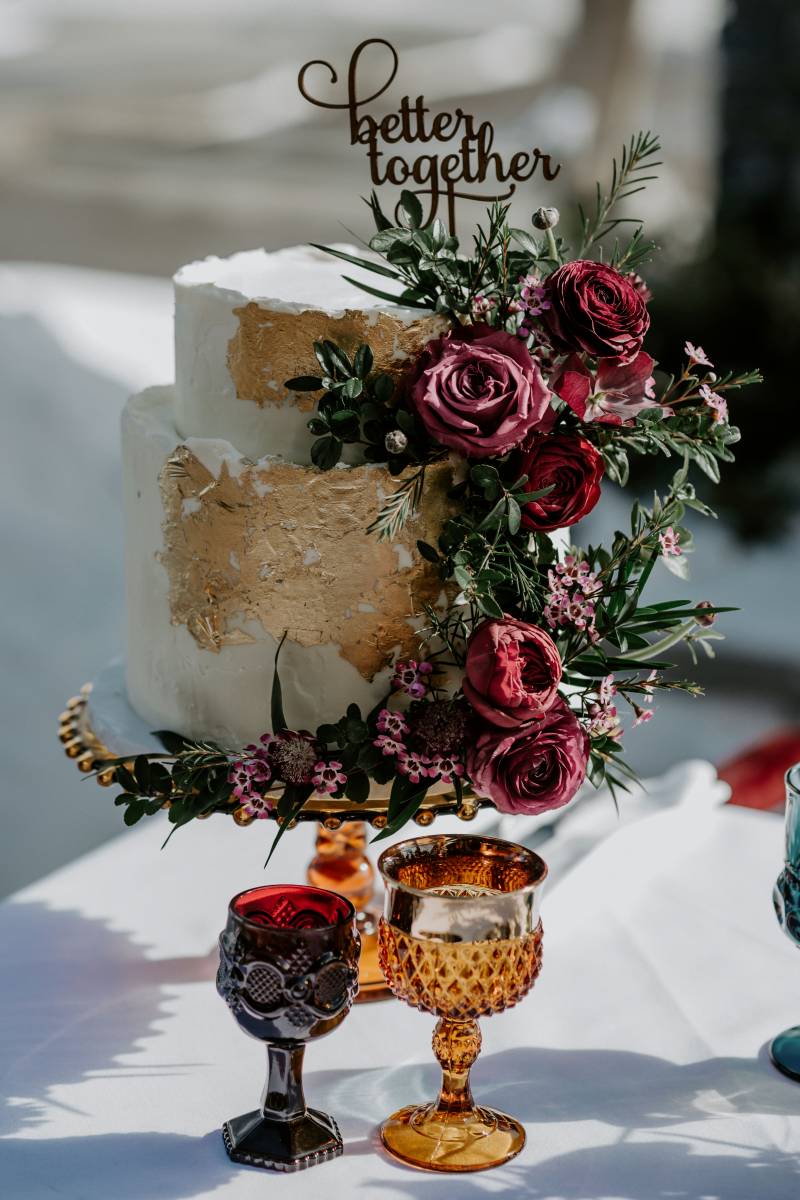 White wedding cake with gold foil on outside and red floral arrangements with gold and red goblets in front 