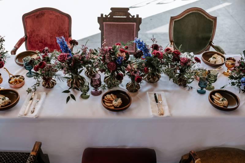 Dark red and blue floral arrangement on white table surrounded by forest green, lavender and red seats