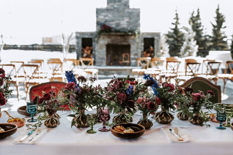 Dark red and blue floral arrangements on white table in front of outdoor stone fireplace 