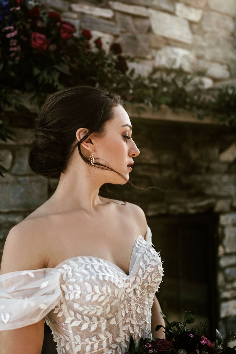 Woman in white lace dress looks down holding dark red bouquet in front of stone fireplace 